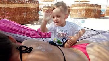 4k视频可爱的小男孩涂防紫外线乳液在母亲背上躺在海边的休息室